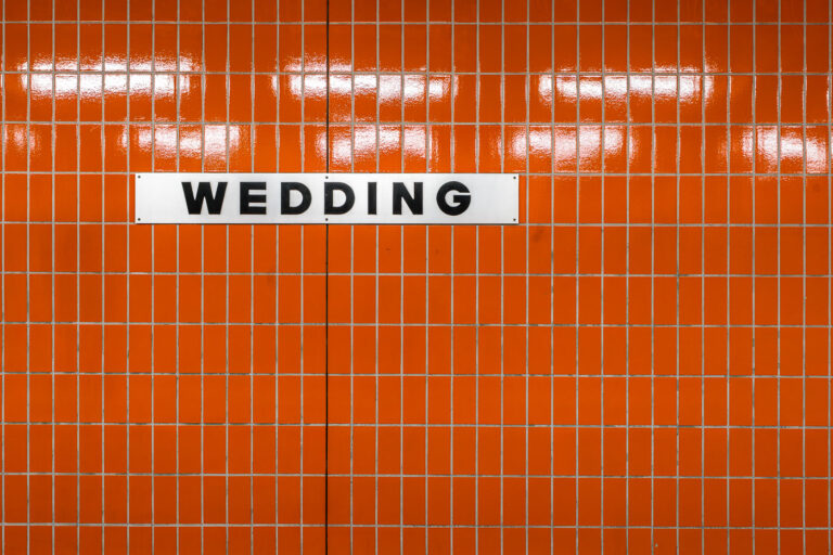 Wedding station sign, Berlin 1 A white station sign reading “Wedding” in black block letters is mounted on an orange, small-rectangle tiled wall inside Wedding station in Berlin, Germany. The station serves the Wedding neighborhood in the Mitte district and functions as an interchange between the U6 U‑Bahn line and the Berlin Ringbahn S‑Bahn. The stark typography and modular tiles reflect the practical design found across much of Berlin’s postwar transit architecture. Operated by BVG for the U‑Bahn and S‑Bahn Berlin GmbH for suburban rail, Wedding is a local hub for commuters moving between the north of the city and central Berlin. No passengers are visible in the frame, emphasizing the graphic clarity of the station’s wayfinding.