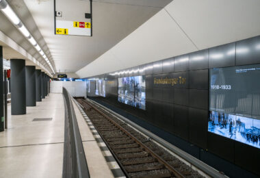 The Brandenburger Tor station on Berlin’s U5 line sits directly beneath Pariser Platz, adjacent to the Brandenburg Gate. The interior features dark composite wall panels inlaid with gold lettering and a series of illuminated displays tracing Berlin’s 20th-century history, including the city’s division and post-reunification development. Originally opened in 2009 as part of the short U55 shuttle between Hauptbahnhof and Bundestag, the station became a through stop in December 2020 when the U5 extension to Alexanderplatz was completed, reconnecting the eastern and western portions of the U-Bahn network after nearly six decades of separation.