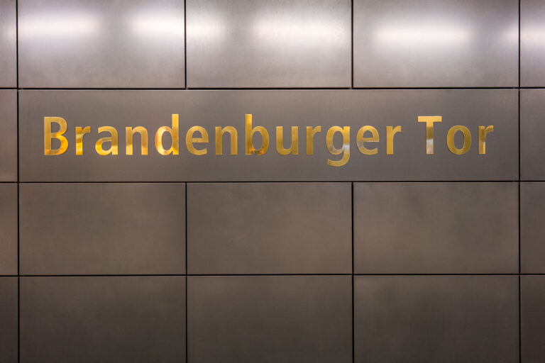 Brandenburger Tor Train Station 2 Gold-lettered signage reading “Brandenburger Tor” is set into a grid of dark, brushed metal panels on the platform wall of Berlin’s Brandenburger Tor station. The underground stop serves as an interchange between the U5 line and the north–south S‑Bahn in the city’s government quarter, steps from the Brandenburg Gate and Pariser Platz. The S‑Bahn station opened in 1936 as Unter den Linden, became a Cold War “ghost station” from 1961 to 1989, and was renamed Brandenburger Tor in 2009. The U‑Bahn platforms, opened in 2009 and fully integrated into the extended U5 in 2020, make the site a key public transit node for commuters and visitors to central Berlin.