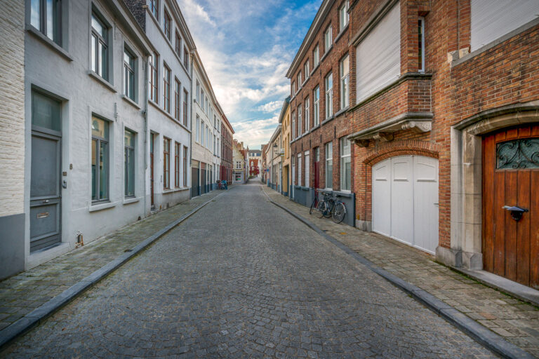 Cobblestone side street in historic Bruges 3 A quiet residential lane in Bruges, Belgium, stretches between rows of brick and plastered townhouses. Cobblestones pave the narrow street, which is lined with ground‑floor doors, shuttered windows, and a white arched garage entrance on the right. Several bicycles are parked against the façades, and a small traffic sign is visible toward the intersection in the distance. The scene sits within Bruges’ historic center, a UNESCO World Heritage site known for its well‑preserved medieval street grid and domestic architecture. Daytime clouds drift over the low‑rise buildings, and no pedestrians or vehicles are present.