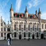 Bruges City Hall (Stadhuis van Brugge), completed in 1421, is one of the oldest and most ornate Gothic town halls in the Low Countries. Located on Burg Square in the heart of Bruges, Belgium, the structure features a richly detailed facade with vertical pinnacles, traceried windows, and over fifty sculpted figures representing biblical and historical figures important to the city’s heritage. The red dormer windows punctuating the steep slate roof are typical of Flemish civic architecture from the period. To the left, the adjoining Renaissance-style Oude Civiele Griffie (Old Civil Registry), built in 1537, complements the Gothic hall with gilded stonework and classical ornamentation. Together, the buildings form a symbolic center of Bruges’ civic identity and remain a key stop for visitors exploring this UNESCO World Heritage city.