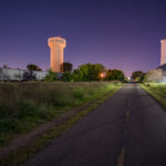 The Cedar Lake Trail passes through the industrial district of St. Louis Park, Minnesota, illuminated here by the community’s water tower and nearby grain elevators. This segment of the trail follows a former railroad right-of-way once used by the Minneapolis & St. Louis Railway, which connected local industries to the region’s freight network. The corridor’s transformation into one of the country’s first bicycle “freeways” in the 1990s marked a major milestone in urban redevelopment and sustainable transportation planning.

Once lined with lumberyards, steel fabricators, and mills, the area surrounding the trail reflects the industrial origins of St. Louis Park—a community that grew rapidly in the early 20th century due to its proximity to both Minneapolis and key rail junctions. The trail today bridges that history, offering cyclists and commuters a route through a landscape where freight trains, utility towers, and legacy industry remain active reminders of the city’s manufacturing past.