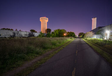 The Cedar Lake Trail passes through the industrial district of St. Louis Park, Minnesota, illuminated here by the community’s water tower and nearby grain elevators. This segment of the trail follows a former railroad right-of-way once used by the Minneapolis & St. Louis Railway, which connected local industries to the region’s freight network. The corridor’s transformation into one of the country’s first bicycle “freeways” in the 1990s marked a major milestone in urban redevelopment and sustainable transportation planning.

Once lined with lumberyards, steel fabricators, and mills, the area surrounding the trail reflects the industrial origins of St. Louis Park—a community that grew rapidly in the early 20th century due to its proximity to both Minneapolis and key rail junctions. The trail today bridges that history, offering cyclists and commuters a route through a landscape where freight trains, utility towers, and legacy industry remain active reminders of the city’s manufacturing past.