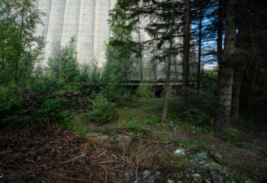Dense vegetation surrounds the concrete base of a decommissioned cooling tower in Charleroi, Belgium. The structure’s massive form rises from a patch of overgrowth and debris, where nature has begun reclaiming the once-industrial site. The cooling tower, part of an abandoned power plant complex, remains a reminder of the region’s post-industrial landscape and transition away from coal-era energy infrastructure.