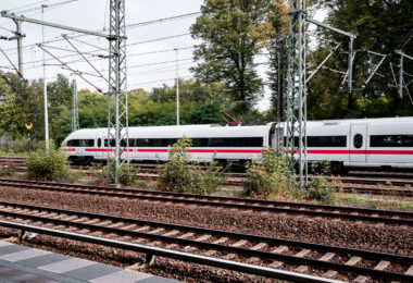 A Deutsche Bahn Intercity-Express (ICE) train moves through the rail network, distinguished by its white body and red horizontal stripe. The train belongs to the ICE 3 or ICE T family, units designed for high-speed operation up to 300 km/h and used on long-distance routes across Germany and neighboring countries. Overhead catenary lines supported by lattice-steel gantries deliver 15 kV AC power, the standard for Germany’s electrified mainlines.

The ICE network, launched in the early 1990s, forms the backbone of long-distance rail travel in the country, linking cities such as Berlin, Frankfurt, Cologne, and Munich. These trains are built for both speed and efficiency, with lightweight aluminum bodies, regenerative braking, and pressure-sealed cabins for tunnel travel. The scene reflects the characteristic organization of German rail infrastructure—multiple tracks, overhead power, and vegetation at the margins—where local and international services share one of Europe’s busiest and most interconnected systems.