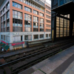 Elevated tracks at Berlin Friedrichstraße station 1 View from the platform at Berlin Friedrichstraße station overlooking Georgenstraße below. The elevated tracks run alongside a mix of modern and postwar office buildings, including the Aerztezentrum at No. 147. Pedestrians and cyclists move through the busy commercial street beneath, while the station’s glass and steel structure reflects the late-day light.