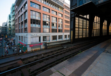 View from the platform at Berlin Friedrichstraße station overlooking Georgenstraße below. The elevated tracks run alongside a mix of modern and postwar office buildings, including the Aerztezentrum at No. 147. Pedestrians and cyclists move through the busy commercial street beneath, while the station’s glass and steel structure reflects the late-day light.
