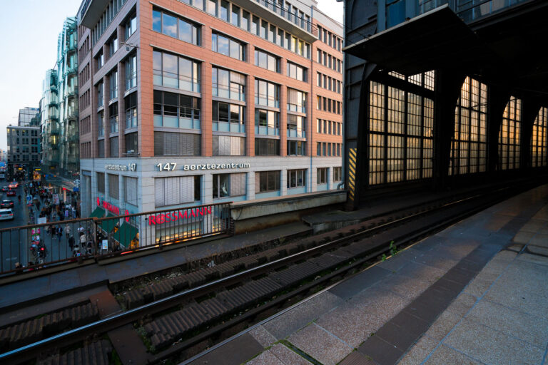 Elevated tracks at Berlin Friedrichstraße station 2 View from the platform at Berlin Friedrichstraße station overlooking Georgenstraße below. The elevated tracks run alongside a mix of modern and postwar office buildings, including the Aerztezentrum at No. 147. Pedestrians and cyclists move through the busy commercial street beneath, while the station’s glass and steel structure reflects the late-day light.