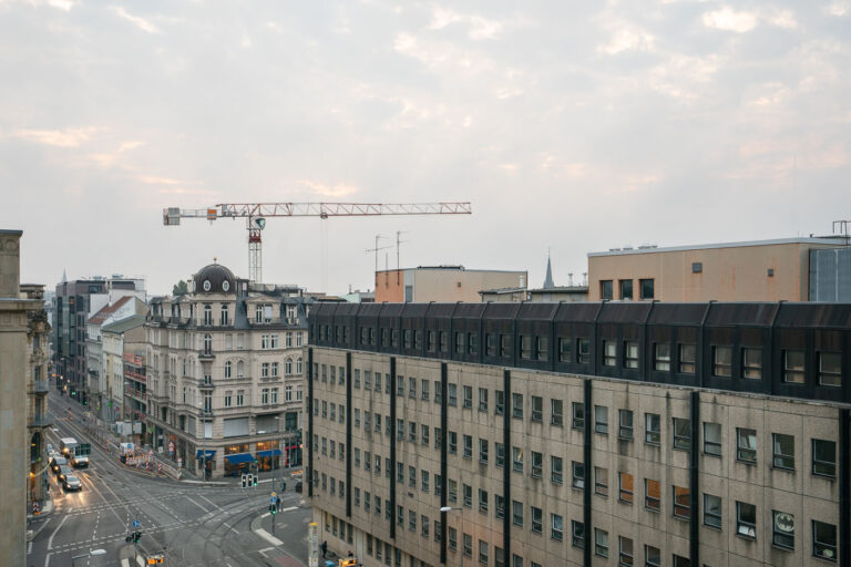 Invalidenstrabe Berlin Germany 6 An early evening view over central Berlin captures the intersection of Friedrichstraße and Dorotheenstraße near the Spittelmarkt area. Traffic lights reflect off the damp pavement as trams and cars navigate the junction, while a construction crane rises above the rooftops—evidence of the city’s ongoing redevelopment and modernization efforts.In the foreground, postwar concrete office buildings contrast with a restored 19th-century corner structure featuring a domed roof and classical detailing. The muted light and overcast sky lend the scene a subdued atmosphere typical of Berlin’s autumn season, illustrating the coexistence of historical architecture, socialist-era infrastructure, and contemporary urban growth.