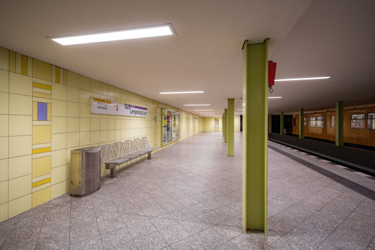 Leopoldplatz U-Bahn Station Platform 2 This view of Leopoldplatz’s lower-level U9 platform highlights the minimalist postwar design typical of 1960s Berlin infrastructure. The use of pale yellow tiles, stainless steel seating, and green-painted support columns was chosen to create a bright, efficient environment for high-traffic commuter service. The station connects to major commercial and residential zones and is part of the BVG modernization program maintaining accessibility and structural updates across Berlin’s U-Bahn network.