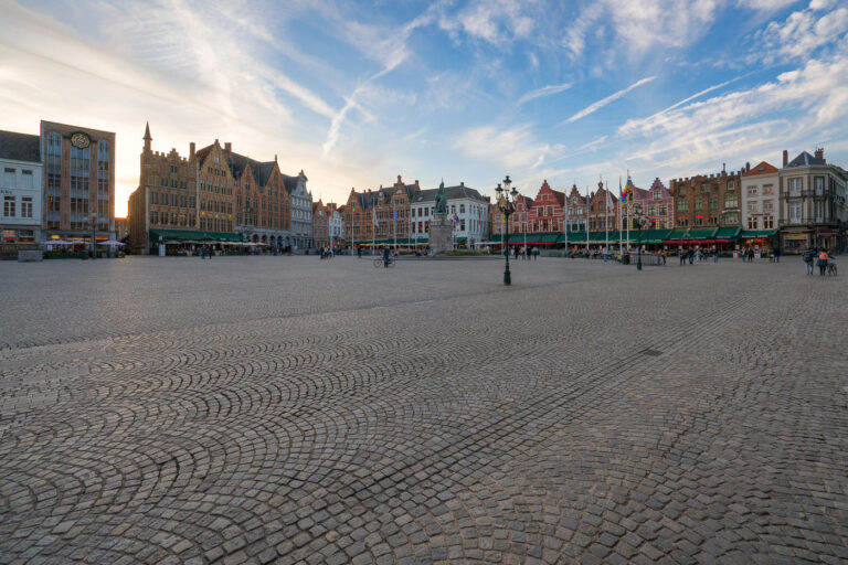 Sunset over the Markt in Bruges 2 Evening on the Markt, Bruges, Belgium. Visitors cross the wide cobblestoned square as sunset light outlines the row of step‑gabled guild houses that front cafés with green awnings. At the center stands the 1887 monument to Jan Breydel and Pieter de Coninck, ringed by flagpoles and a cast‑iron lamppost. The scene captures a typical early evening in Bruges’ historic core, a UNESCO‑listed medieval city where the Markt has hosted trade and civic life for centuries. Contrails and thin clouds streak the sky while small groups linger near bicycles and restaurant terraces.
Keywords
- Activities: strolling, sightseeing, cycling, dining
- Buildings: step-gabled guild houses, cafés, monument plinths
- Location: Bruges, West Flanders, Belgium; Market Square (Markt)
- Objects: cobblestones, lamppost, flagpoles, café awnings, clock
- People: tourists, locals, pedestrians
- Moods: calm, unhurried, early evening
- Sceneries: historic town square, open plaza
- Texts: storefront signage, building numbers (unreadable at distance)
- Companies: local cafés and restaurants (unidentified)
- Weather: clear with high clouds at sunset
- Plants: none visible
- Animals: none visible
- Vehicles: bicycles