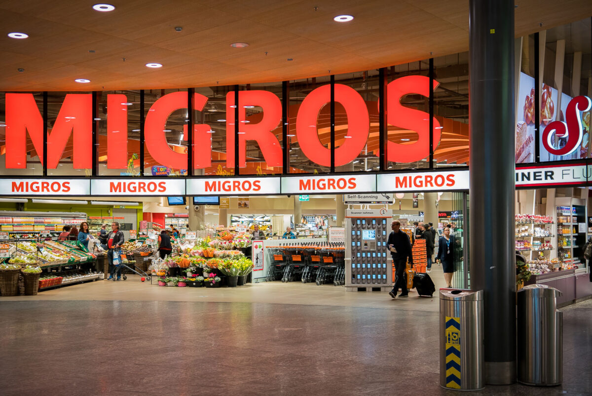 Entrance to a Migros supermarket, a major Swiss retail chain. Shoppers move in and out with carts and luggage, while displays of flowers and fresh produce line the front of the store. Large illuminated signage with the store’s name dominates the facade.