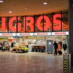 Entrance to a Migros supermarket, a major Swiss retail chain. Shoppers move in and out with carts and luggage, while displays of flowers and fresh produce line the front of the store. Large illuminated signage with the store’s name dominates the facade.