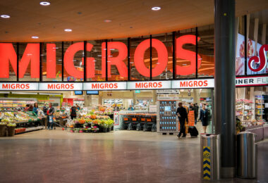 Entrance to a Migros supermarket, a major Swiss retail chain. Shoppers move in and out with carts and luggage, while displays of flowers and fresh produce line the front of the store. Large illuminated signage with the store’s name dominates the facade.