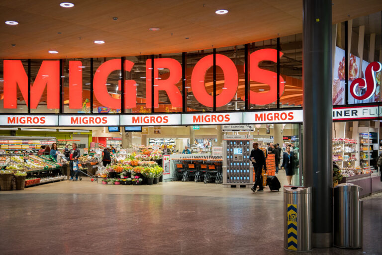 Migros Supermarket in Zurich, Switzerland 4 The entrance of a Migros supermarket in Zurich, Switzerland, is shown in this photo. The large, illuminated red sign spelling 'MIGROS' dominates the storefront, with smaller signs repeating the name above the entrance. Inside, shoppers are seen browsing various sections, including fresh produce and flowers, with shopping carts and self-scanning kiosks visible. The setting appears to be during daytime or early evening, with a lively atmosphere typical of a busy retail location in Switzerland.