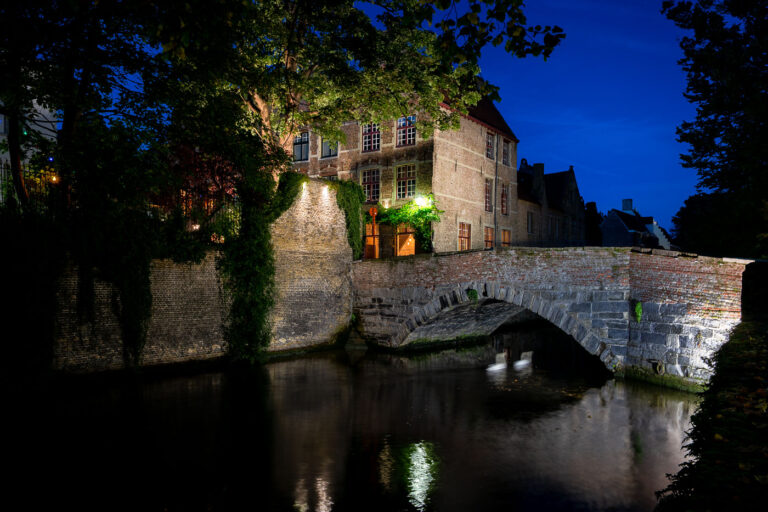 Night view of a historic bridge over the River Reie in Bruges 4 Medieval bridge on the Reie, Bruges at dusk. A single-arch stone and brick bridge spans a quiet bend of the River Reie in Bruges, Belgium, its parapet and adjoining quay lit by warm lamps that reflect on the canal. Behind the bridge, ivy climbs weathered brick facades and gabled townhouses typical of the city’s historic core, a UNESCO World Heritage site. Leafy trees frame the scene, and the waterway shows faint ripples but no pedestrians or boats are visible. The location sits along one of Bruges’ centuries-old canals that once supported trade and still anchor the city’s tourism and daily life. Keywords — Activities: evening strolls, sightseeing; Buildings: historic townhouses, canal quay; Location: Bruges, Belgium, River Reie, historic center; Objects: streetlights, ivy-covered walls, windows; People: none visible; Moods: tranquil, contemplative; Sceneries: canal, old bridge, reflections; Texts: none; Companies: none; Weather: clear evening; Plants: trees, ivy; Animals: none visible; Vehicles: none visible.