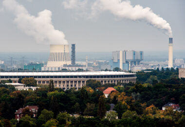 The Olympiastadion in Berlin stands in the foreground, framed by the dense tree cover of the Westend district. Behind it, the cooling towers and chimneys of the Reuter power plants dominate the skyline — part of Berlin’s long-standing energy infrastructure along the Spree River. The large natural-draft cooling tower at left belongs to the Reuter West plant, commissioned in the late 1980s and operated by Vattenfall, while the taller stack at right is part of the older Reuter facility dating back to 1930, later converted to modern combined-heat-and-power operation.

The contrast between the stone colonnades of the 1936 Olympic Stadium and the industrial silhouettes in the distance captures the layered character of Berlin’s western edge — where historic architecture, post-war redevelopment, and ongoing energy production coexist within a few kilometers. The area remains one of the city’s key power-generation corridors, supplying electricity and district heating to much of central and western Berlin.