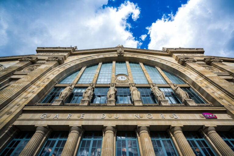 Beaux-Arts entrance of Paris’s Gare du Nord 1 Gare du Nord’s monumental Beaux‑Arts facade is pictured from street level in the 10th arrondissement of Paris, France. The stone frontage features tall arched windows, a central clock, and a row of allegorical statues that represent major European cities historically served by the station’s routes. The inscription “GARE DU NORD” is set above fluted columns, and a magenta SNCF logo marks the national rail operator; cumulus clouds break over a blue sky above the cornice. Opened in the 1860s and designed by architect Jacques-Ignace Hittorff, Paris-Nord is among Europe’s busiest rail hubs, linking regional Transilien lines, RER services, the Paris Métro, and international trains including the Eurostar to London. The station has long served as a gateway for commuters and cross‑border travelers, reflecting the growth of rail travel in northern Europe since the 19th century.