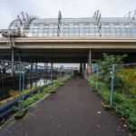 A narrow, fenced pathway runs beneath the towering structure of Berlin Hauptbahnhof — Germany’s largest and most complex railway station — where elevated train lines stretch above the Spree River. The gleaming glass panels and steel framework of the upper platforms contrast sharply with the damp pavement and industrial underpass below. Puddles glisten from a recent rain, while construction barriers and wild greenery frame the scene, hinting at the city’s perpetual state of transformation. Built as a symbol of reunified Berlin, the Hauptbahnhof connects north to south, east to west — a convergence of architecture, engineering, and history that mirrors Berlin’s layered identity. The stillness here beneath the trains creates a hidden perspective on one of Europe’s busiest transport hubs, where modern motion meets quiet decay.
