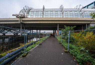 A narrow, fenced pathway runs beneath the towering structure of Berlin Hauptbahnhof — Germany’s largest and most complex railway station — where elevated train lines stretch above the Spree River. The gleaming glass panels and steel framework of the upper platforms contrast sharply with the damp pavement and industrial underpass below. Puddles glisten from a recent rain, while construction barriers and wild greenery frame the scene, hinting at the city’s perpetual state of transformation. Built as a symbol of reunified Berlin, the Hauptbahnhof connects north to south, east to west — a convergence of architecture, engineering, and history that mirrors Berlin’s layered identity. The stillness here beneath the trains creates a hidden perspective on one of Europe’s busiest transport hubs, where modern motion meets quiet decay.