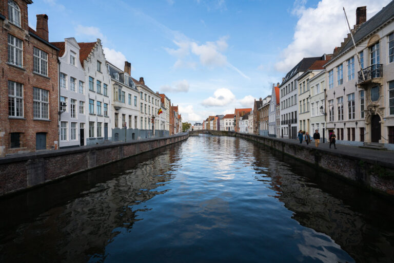 Historic canal houses along the River Reie in Bruges 2 River Reie canal through Bruges’ historic center. Rows of brick and whitewashed townhouses with stepped gables line both banks of the River Reie in Bruges, Belgium. A Belgian tricolor flag hangs from one façade, and wrought-iron street lamps and balconies frame tall windows along the quay. Several pedestrians walk the right-hand embankment as calm water reflects the buildings and a low arched bridge visible in the distance. The scene is photographed in daylight under a partly cloudy sky within the UNESCO-listed medieval core known for its preserved canal network. Keywords — Activities: walking, sightseeing; Buildings: gabled townhouses, historic façades; Location: Bruges, Belgium, River Reie, historic center; Objects: Belgian flag, street lamps, balconies, canal walls; People: pedestrians, tourists; Moods: calm, reflective; Sceneries: canal, waterfront, bridge; Texts: none visible; Companies: none; Weather: partly cloudy, fair; Plants: sparse greenery on quay; Animals: none; Vehicles: none.