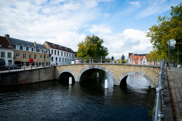 Arched bridge over the River Reie, Bruges 2 Pedestrians cross a three-arched brick bridge over a branch of the River Reie in Bruges, Belgium. Historic row houses with red-tiled roofs and a mix of gabled and flat façades line the canal, while bollards and railings mark the waterway and walkway. Trees showing early autumn color frame the scene, and a partly cloudy sky brightens the medieval streetscape. The canal network, once the commercial lifeline of the city, is part of the UNESCO-listed Historic Centre of Brugge and remains a focal point for daily life and tourism.