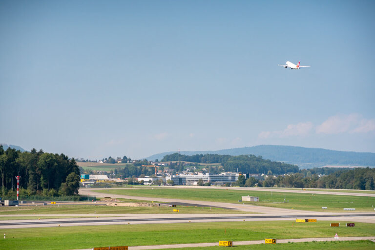Departure from Zurich Airport Runway 3 A Swiss International Air Lines jet climbs into the clear summer sky after takeoff from Zurich Airport (Flughafen Zürich), leaving behind the rolling green hills of Kloten in the background. The well-marked taxiways and precise runway layout reflect Switzerland’s meticulous approach to aviation infrastructure. Beyond the airport, clusters of small homes and industrial buildings blend into the forested landscape — a typical Swiss juxtaposition of modern efficiency and natural calm.