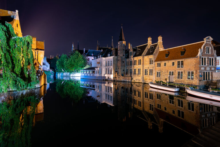 Night Reflections on Bruges’ River Reie 1 Nighttime reflections line a bend of the River Reie in Bruges, Belgium, as historic brick guild houses and stepped-gable facades mirror perfectly on the still canal. Two white tour boats sit moored for the night along the quay, while a vine-draped wall and a waterside restaurant pavilion are illuminated across the water. Church spires rise in silhouette behind the row of houses, anchoring the skyline of the medieval core. Bruges’ historic center, a UNESCO World Heritage Site, is known for its preserved canals and architecture that draw heavy daytime tourism; after hours, the waterways fall quiet except for lights from hotels and eateries along the quay.