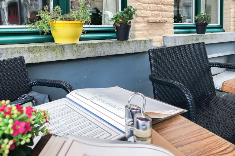 Outdoor Café Table in Bruges 3 A small café table set along a cobblestone street in Bruges, Belgium, with a menu laid open beside salt and pepper shakers and woven black chairs. The table sits beneath pastel-colored brick façades and flower pots on stone ledges—typical of the city’s historic core where cafés blend seamlessly into centuries-old townhouses. Outdoor seating like this is common throughout Bruges’ quieter lanes, offering a relaxed view of daily life amid the preserved medieval architecture.