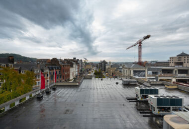 Looking along Rue de Fer toward Namur’s main station in Wallonia, Belgium. Historic brick and limestone façades line the street, with the HEMA storefront sign visible among local shops and cafés. Buses and pedestrians move through the late afternoon traffic on a damp autumn day, reflecting the city’s mix of preserved 19th-century architecture and contemporary commercial life near the railway hub.