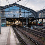 Berlin Friedrichstraße train station 2 Berlin Friedrichstraße station in the early evening, with an S-Bahn BR 481 series train stopped under the steel and glass canopy. The curved elevated tracks lead into the main hall, a design characteristic of the city’s prewar rail architecture. Overhead wires and support structures frame the scene, while a few commuters move along the quiet platform as daylight fades into artificial light.