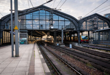 Berlin Friedrichstraße station in the early evening, with an S-Bahn BR 481 series train stopped under the steel and glass canopy. The curved elevated tracks lead into the main hall, a design characteristic of the city’s prewar rail architecture. Overhead wires and support structures frame the scene, while a few commuters move along the quiet platform as daylight fades into artificial light.