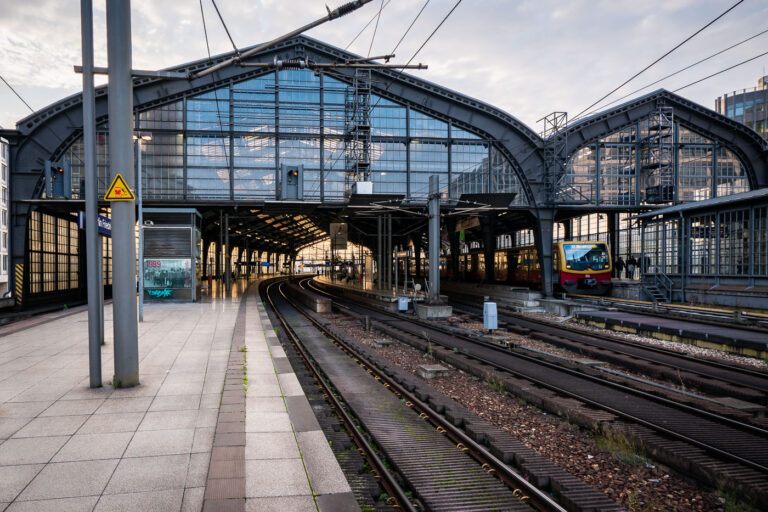 Berlin Friedrichstraße train station 2 Berlin Friedrichstraße station in the early evening, with an S-Bahn BR 481 series train stopped under the steel and glass canopy. The curved elevated tracks lead into the main hall, a design characteristic of the city’s prewar rail architecture. Overhead wires and support structures frame the scene, while a few commuters move along the quiet platform as daylight fades into artificial light.