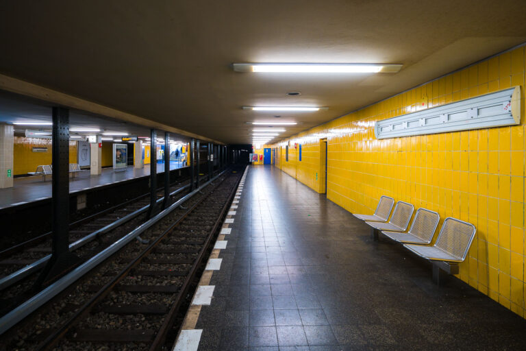 Seestrabe U-Bahn Station in Berlin 1 Seestraße U-Bahn platforms in Berlin’s Wedding district sit empty, their yellow-tiled walls and metal benches illuminated by ceiling fixtures. Twin side platforms flank two tracks, with black steel columns, track ballast, tactile edging, stairways, and signage reading “Seestraße” visible across the way. The station serves Line U6 of the Berliner Verkehrsbetriebe (BVG) network, a north–south route linking Tegel with the city center and beyond. Opened in 1923 on the former Line C, Seestraße reflects the interwar expansion of Berlin’s underground and remains a neighborhood transit hub connecting residents to tram and bus services at street level.