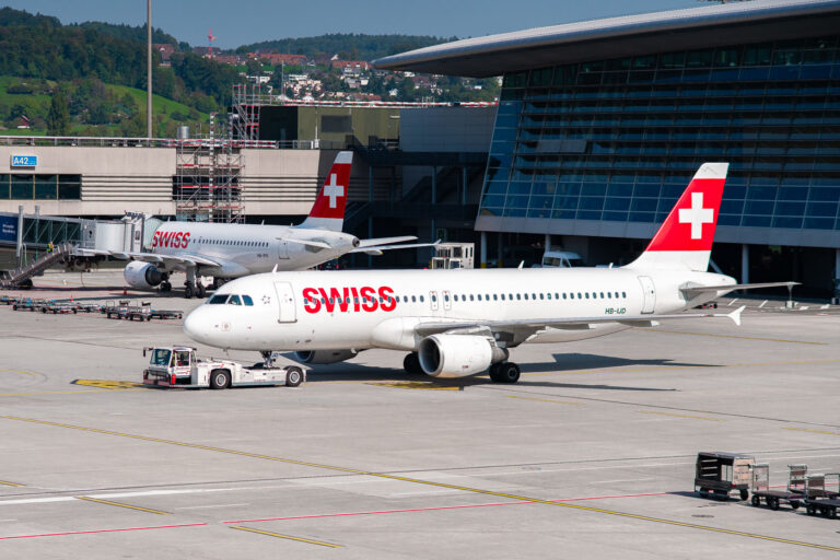 Swiss A320 Fleet at Zurich Airport 1 Two Swiss International Air Lines Airbus A320 aircraft sit on the ramp at Zurich Airport, their red and white tails bearing the iconic Swiss cross. The foreground jet, registration HB-IJD, is being towed toward its gate while another prepares for boarding nearby. Behind them, the glass façade of the terminal reflects the surrounding hills of Kloten, a reminder of Switzerland’s seamless integration of precision engineering and alpine landscape. The scene captures the calm efficiency of Swiss aviation — clean, orderly, and unmistakably national in character.