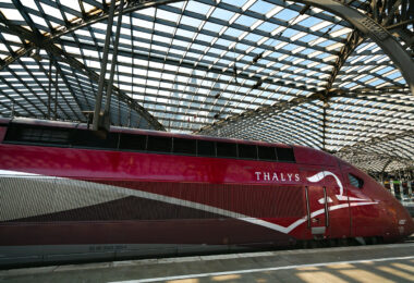 A Thalys PBKA high-speed train waits at Köln Hauptbahnhof beneath the station’s large arched glass canopy. The streamlined red train, built by Alstom and operated on international routes between Germany, Belgium, France, and the Netherlands, reflects the late-afternoon light along its metallic bodywork. Overhead, the steel lattice roof filters the daylight across the platforms, and through the patterned glass, the spires of Cologne Cathedral rise faintly in the background—a visual reminder of the station’s central location beside one of Europe’s most recognizable landmarks.