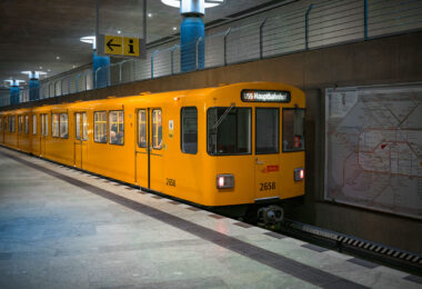 A bright yellow BVG U-Bahn train marked “U55 Hauptbahnhof” stands at the underground platform of Berlin Hauptbahnhof. An information sign with an arrow hangs above the platform, while a large network map of the S- and U-Bahn lines is mounted on the concrete wall to the right. Through the windows, a few seated passengers are visible inside the multi-door carriage numbered 2658. The U55 operated as a short shuttle between Berlin Hauptbahnhof and Brandenburger Tor, opened in 2009 to link the new central station with the government quarter. In December 2020 the shuttle was integrated into the extended U5 line, a major step in Berlin’s east‑west metro connection.