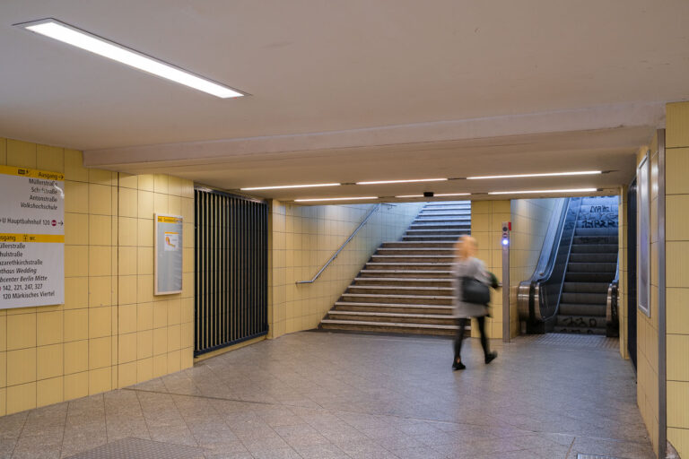 Leopoldplatz U-Bahn Station in Berlin 3 An underground passageway connects the platforms of Wedding U-Bahn station to its street-level exits on Müllerstraße in Berlin’s Mitte district. The corridor is tiled in light yellow ceramic, part of the BVG’s standardized postwar design used throughout many West Berlin stations rebuilt or renovated in the 1960s and 1970s.Directional signage guides passengers toward exits, connecting bus routes, and public facilities, while an escalator and stairway lead to the main entrance. The faint graffiti on the walls and motion blur of a passing commuter emphasize the station’s everyday function within Berlin’s public transport network. Wedding station serves both the U6 line and the S-Bahn Ring, forming a key interchange point in the city’s northern transit system.