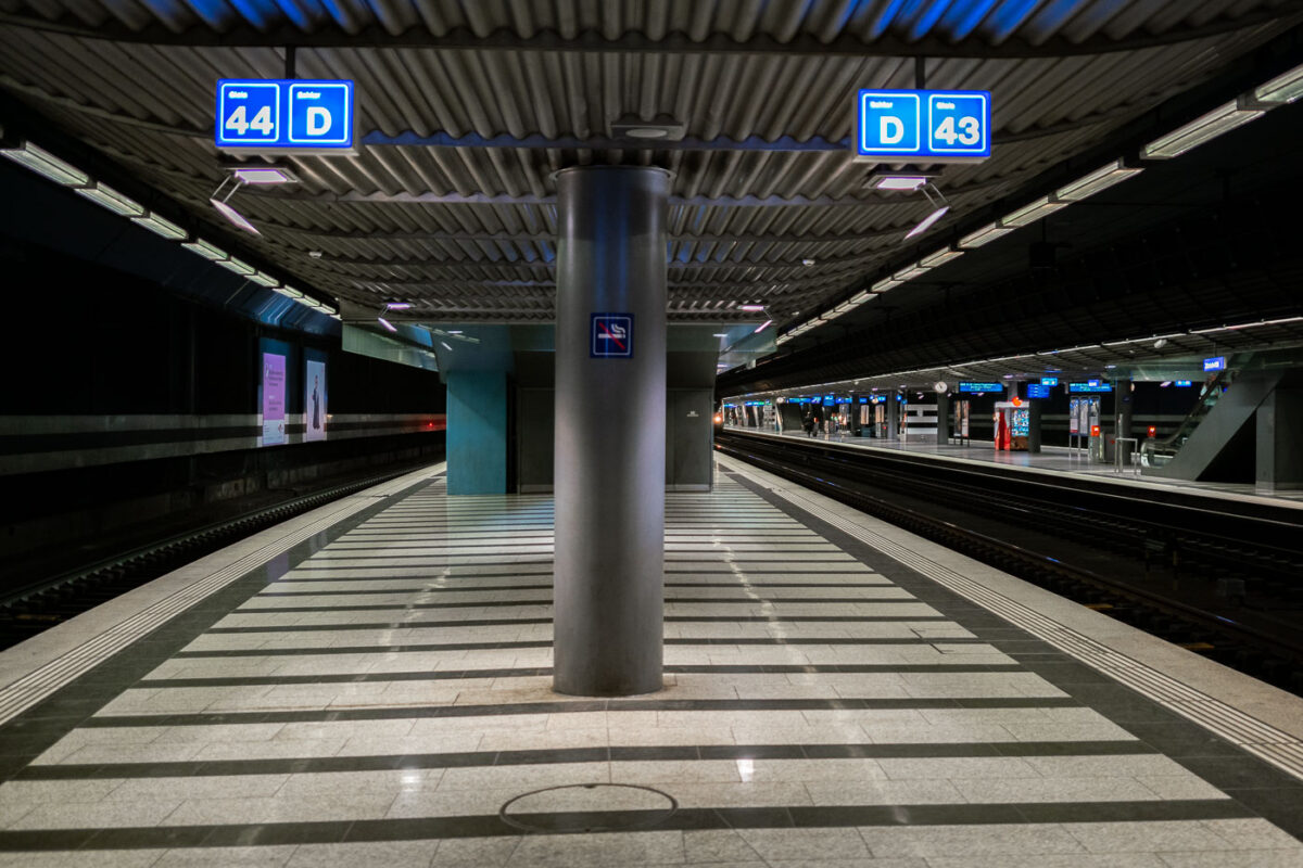 An underground train platform at Zürich Airport station, with tracks 43 and 44 visible. The station features striped tile flooring, overhead signage, and modern lighting. Few passengers are present, and escalators can be seen in the background leading to the upper levels.