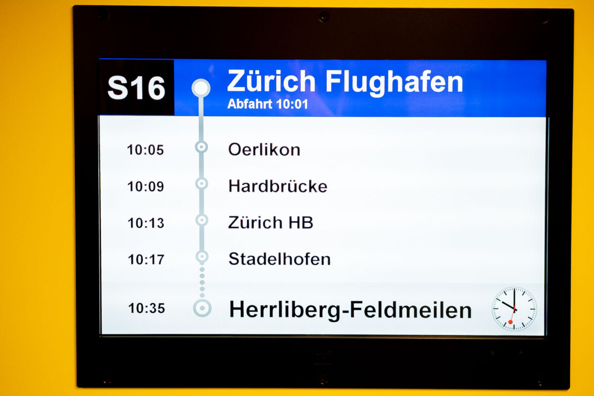 A digital departure board at Zürich Airport displays the S16 train schedule. The train departs at 10:01 and makes stops at Oerlikon, Hardbrücke, Zürich Hauptbahnhof, and Stadelhofen before arriving in Herrliberg-Feldmeilen at 10:35. A Swiss railway clock is shown in the lower right corner.