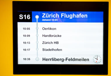 A digital departure board at Zürich Airport displays the S16 train schedule. The train departs at 10:01 and makes stops at Oerlikon, Hardbrücke, Zürich Hauptbahnhof, and Stadelhofen before arriving in Herrliberg-Feldmeilen at 10:35. A Swiss railway clock is shown in the lower right corner.