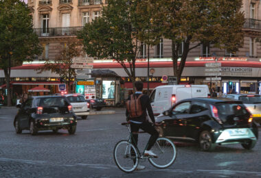 A cyclist waits at an intersection in Paris as evening traffic moves through the roundabout. Street signs point toward Place de Wagram, Asnières, and the périphérique. In the background, the brasserie “Le Lorraine” and Haussmann-style buildings frame the corner of the busy street.