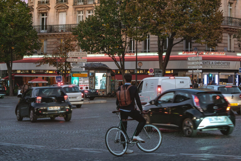Cyclist at Place des Ternes, Paris, Evening Traffic 3 A cyclist navigates evening traffic at Place des Ternes in Paris, a busy roundabout bordering the 8th and 17th arrondissements. The area is characterized by Haussmannian architecture, as seen in the facade of Brasserie La Lorraine, and serves as a key transit point, with signs indicating routes to Porte d’Asnières, the Périphérique, Place de Wagram, and Porte de Clichy. This location, near the Arc de Triomphe corridor, experiences significant rush hour activity, with vehicles and pedestrians utilizing the cobblestone streets and surrounding commercial establishments.