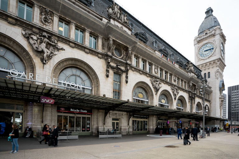 Gare de Lyon in Paris, France 2 The Gare de Lyon train station in Paris, France, is depicted in this photograph taken during daytime. The historic building features ornate architectural details, including sculptures and decorative stonework, with a prominent clock tower displaying the time. The station's entrance is visible, with signs indicating SNCF and other services, and people are seen walking and waiting outside. The scene captures the bustling activity typical of a major transportation hub in the city.