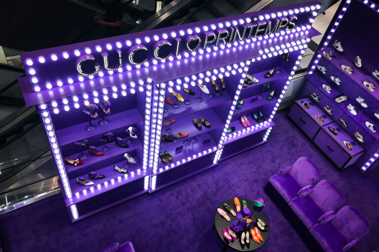 Gucci x Printemps Pop-Up, Paris (2017) 3 A vibrant Gucci x Printemps display inside Paris’s historic Printemps department store, featuring illuminated shelving and an array of designer footwear. The installation’s electric purple décor and marquee-style lighting reflect the brand’s maximalist creative direction under Alessandro Michele during the mid-2010s, a period marked by bold colors and theatrical presentation.
Printemps, founded in 1865, has long been a landmark of Parisian retail innovation—among the first to introduce electric lighting and glass-domed architecture in department stores. Its collaborations with luxury brands like Gucci blend heritage and modernity, cementing its role as both a shopping destination and a showcase for contemporary design trends in the city’s fashion district along Boulevard Haussmann.