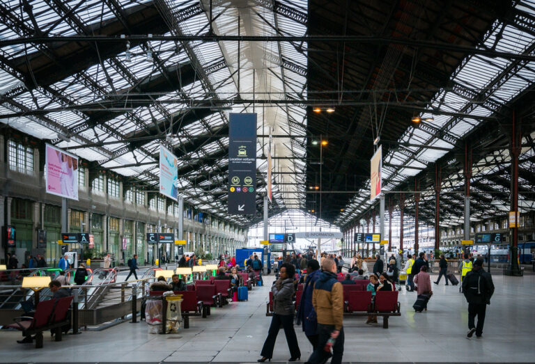 Midday bustle at Paris Gare de Lyon concourse 1 Passengers wait and walk through the main concourse of Gare de Lyon in Paris, France, a major rail hub located in the 12th arrondissement. Rows of red seating, baggage trolleys, and electronic boards marked with concourse letters C through G line the iron-and-glass train hall, while SNCF trains are visible at the platforms to the right. Overhead signage points to “Information,” car rental, and connections to Paris Métro lines 1 and 14 and the RER A and D. The station, opened for the 1900 Exposition Universelle, serves high-speed TGV and regional services toward southeastern France and international destinations, making it one of the city’s busiest gateways.