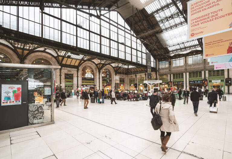 Midday flow in the concourse of Paris Gare de Lyon 4 Passengers cross the main concourse of Paris Gare de Lyon, 12th arrondissement, France, near coordinates. Under the iron-and-glass train shed, travelers wheel suitcases past an information point, rows of red seats, and storefronts with green-trimmed windows. Large banners in French promoting a music-streaming service hang from the rafters, while overhead signs direct people toward Hall 2 and other platforms. The Beaux-Arts arches and clerestory windows, part of a station complex inaugurated for the 1900 Exposition Universelle, frame the busy interior. Gare de Lyon is one of Paris’s principal rail hubs, serving TGV and regional lines to southeastern France and beyond, as well as RER and Métro connections for city transit.
