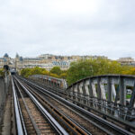 Paris Metro Line 6 crosses the Seine on the Pont de Bir-Hakeim, a two-level bridge completed in 1905 that combines roadway, pedestrian walkway, and elevated railway. The steel viaduct, designed to support one of the city’s earliest elevated metro lines, features riveted beams typical of early 20th-century engineering. In the distance is the Passy district in the 16th arrondissement, known for its Haussmann-era architecture and ornate façades. This elevated section of Line 6 was intentionally built above ground to provide air circulation for the early steam-powered trains, making it one of the few Paris metro routes that offer open views of the city’s urban landscape.