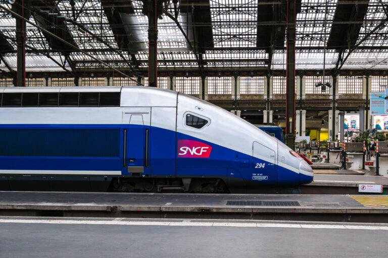 TGV at Gare de Lyon, Paris 3 SNCF high‑speed service at Paris’s Gare de Lyon. A blue‑and‑silver TGV unit marked with the SNCF logo and the number 294 stands at a platform beneath the station’s iron-and-glass train shed at Gare de Lyon in Paris, France. The streamlined nose of the train faces a row of barriers and signage, while maintenance equipment and timetable boards are visible in the concourse behind. Open work bays, columns, and tall windows define the historic terminal architecture, one of the capital’s main gateways to southeastern France. Operated by the national rail company SNCF, Gare de Lyon handles frequent high‑speed services toward Lyon, Marseille, the French Riviera, and cross‑border connections to Switzerland.