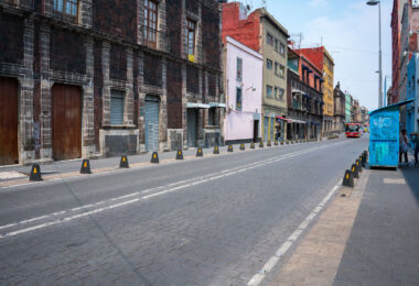 Calle República de Uruguay in the heart of Mexico City’s Centro Histórico, lined with colonial-era stone façades and early 20th-century buildings showing layers of renovation and age. The street, paved with basalt cobblestones, reflects the city’s dense architectural history where 18th-century walls coexist with modern storefronts and utilities. The corridor connects the Zócalo area with Eje Central, serving as one of the oldest commercial arteries in the capital.